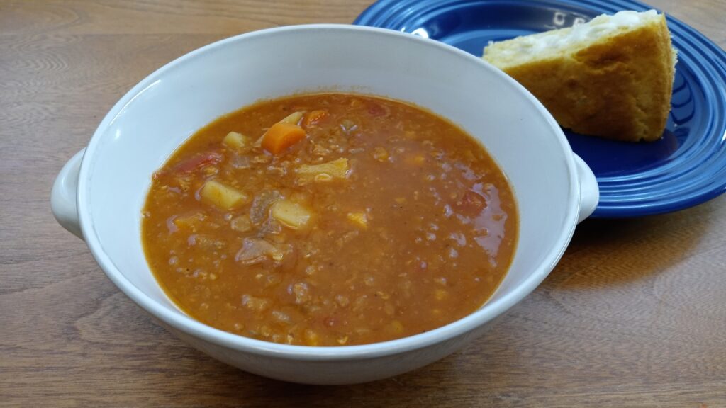 A bowl of spicy winter vegetable soup with corn bread in the background.